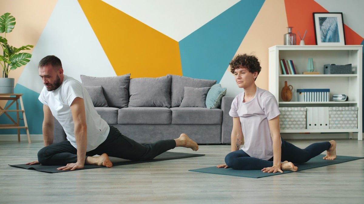 Young couple man and woman are doing yoga at home exercising together on mats practising asanas in apartment moving simultaneously. Youth and sports concept.